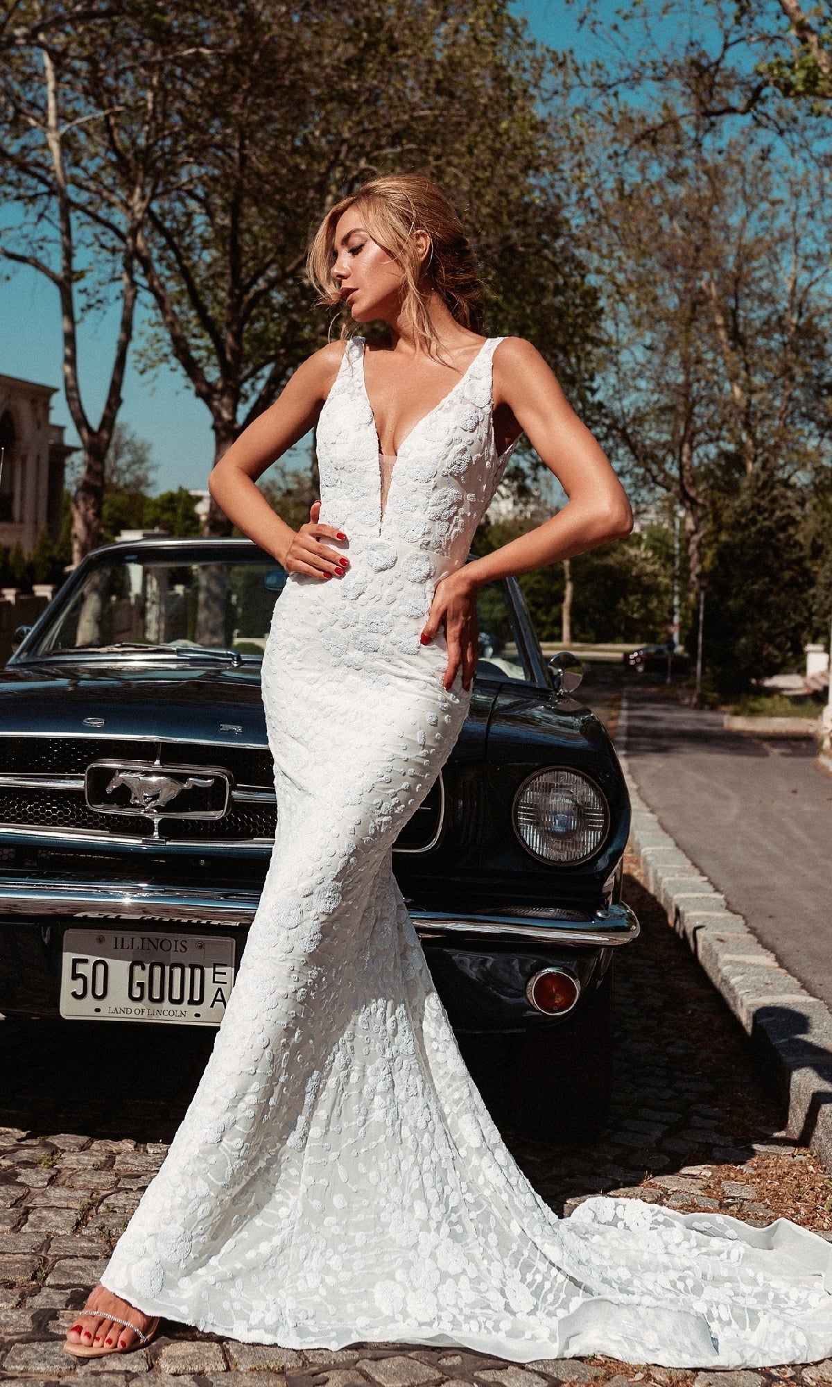 A woman in a dazzling Long White Mermaid Bridal Gown: Tina Holly BB011 poses before a classic black Ford Mustang on a sunny day.